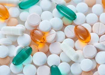 A variety of pills and capsules of different colors and shapes, including white, orange, and green tablets, arranged on a clear surface.