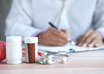 A doctor in a white coat writes on a sheet of paper while a stethoscope and medication bottles are visible on the table.