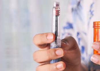 One hand holds an injection device, showing a needle and dose indicator, while the other hand holds an orange container of medication.