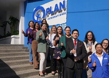 Group of people posing on stairs in front of a blue wall with the Plan International logo, showing thumbs up.