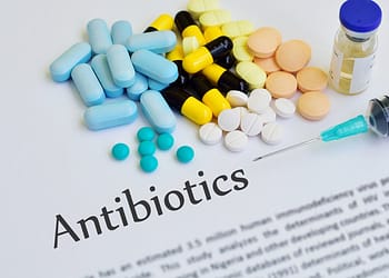 A variety of medications, including pills of different colors and shapes, a syringe and a bottle, on a document that says "Antibiotics."