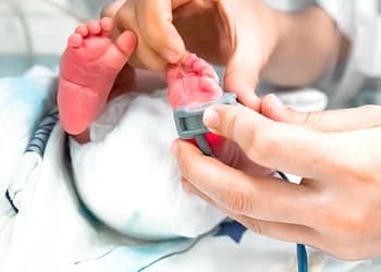 A baby with tiny feet is being examined by a hand placing a monitoring device on one of its feet, while it rests on a surface fitted with medical sheets.