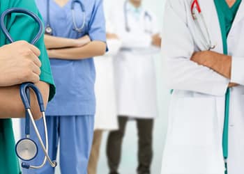 Group of health professionals, with a doctor in a white coat and others in colorful uniforms, some with stethoscopes.