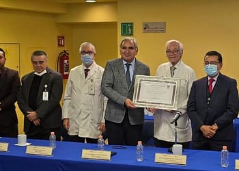 Group of doctors at an event, posing with a framed certificate. They are all wearing masks and standing next to a table with bottles of water. There is a background of yellow walls and decorations.