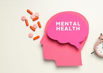 Pink silhouette of a head with a speech bubble that says "MENTAL HEALTH", accompanied by colored pills and a pink alarm clock.