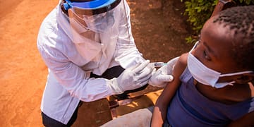 A health care professional wearing protective gear administering a vaccine to a child sitting in a chair outdoors.