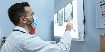 A doctor in a white coat and face mask is pointing at X-ray images under an examination light in a medical office.