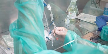 A hand in a clinical setting holds several tubes and syringes, surrounded by laboratory equipment and personal protective equipment, with a background of metallic surfaces and visible pharmaceuticals.
