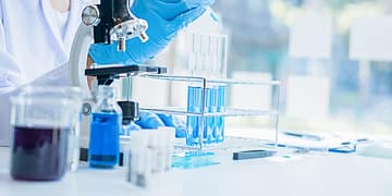 A scientist in a laboratory uses a pipette to add liquid to test tubes containing blue solutions, while a microscope and other laboratory instruments are visible on the table.