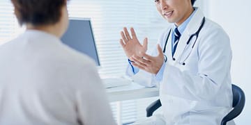 A dark-haired doctor, wearing a white coat and a stethoscope around his neck, is having an animated conversation with a patient in a modern doctor's office setting, with a computer and illuminated windows in the background.