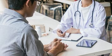 A man sitting opposite a doctor wearing a white coat and stethoscope in an office with a table, a tablet, and several medication bottles visible. The doctor appears to be explaining something to the patient.