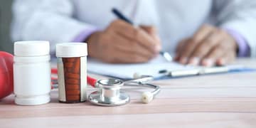 A doctor in a white coat writes on a sheet of paper while a stethoscope and medication bottles are visible on the table.