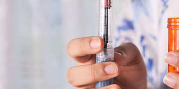 One hand holds an injection device, showing a needle and dose indicator, while the other hand holds an orange container of medication.