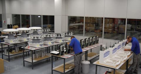 Two workers assemble equipment in a workshop, surrounded by tables with components and work materials. In the background, several complete units are seen ready.