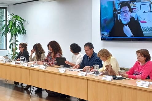 Meeting with participants seated around a long table, some with electronic devices, while a speaker appears on video on a large screen. In the background, there is a decorative plant.