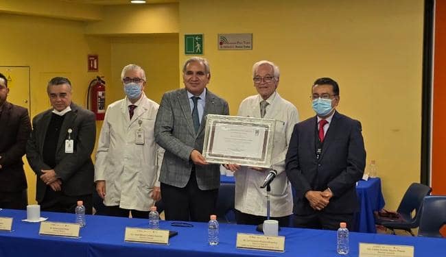 Group of doctors at an event, posing with a framed certificate. They are all wearing masks and standing next to a table with bottles of water. There is a background of yellow walls and decorations.