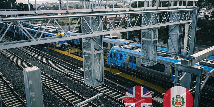 An aerial view of a train station with tracks and parked blue trains, surrounded by an urban environment and metal support structures.