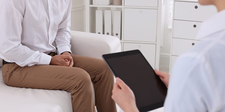 Person sitting on a sofa with hands on legs, while another person holds a tablet, in an office or consulting room environment.