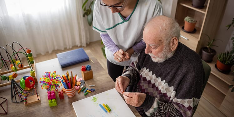 A young woman is helping an elderly man at a table, where there are art materials and toys. The old man, with a gray beard, is concentrating on manipulating an object, while the woman, in a white T-shirt with details, watches attentively. In the background there are plants and colorful toys.