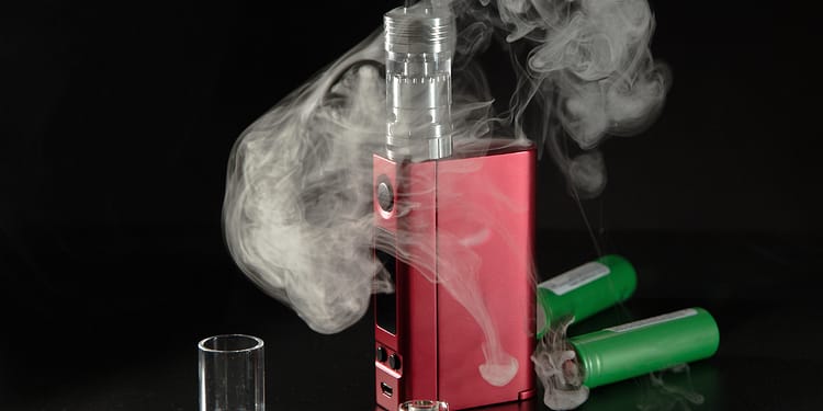 A red vaping device emitting vapor, with two green batteries in the background and a glass cylinder next to it.
