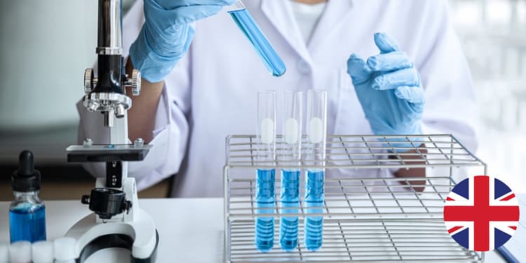 A person in a lab coat and blue gloves holds a test tube with blue liquid, while there are other test tubes in a holder and a microscope on the table.