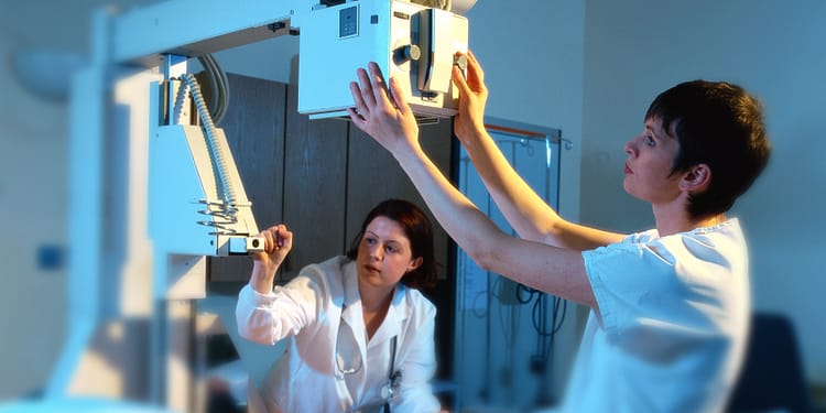Two healthcare professionals in a medical setting, adjusting X-ray equipment, while one of them watches closely.