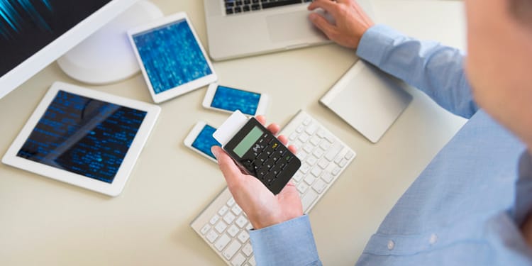 A person holding a calculator in one hand while working at a desk full of electronic devices including a monitor, laptop, tablets and mobile phones.