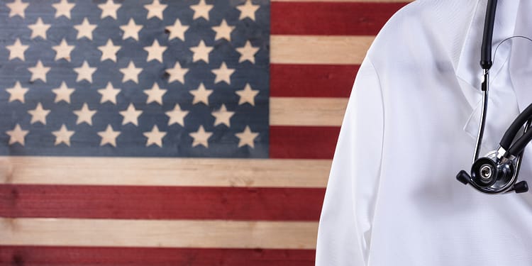 A stethoscope rests on a white coat, in front of a background of the American flag.
