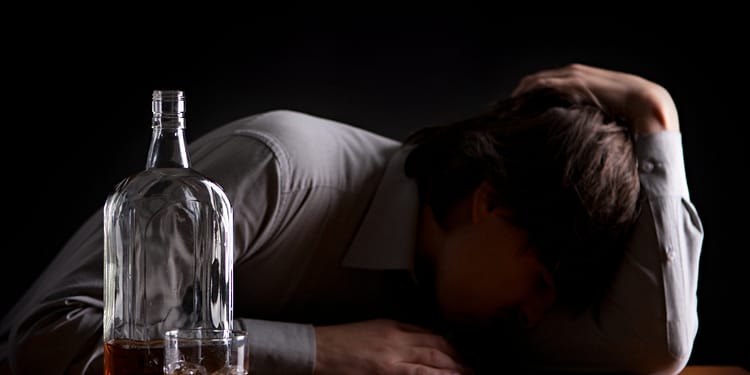 Man in a grey shirt leaning on a wooden table, head down, next to a bottle of liquor and a glass with a drink. Dark background.