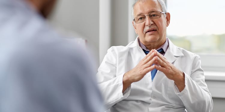 A middle-aged doctor wearing glasses and a white coat sits in front of a patient while talking and gesturing with his hands in a bright, modern doctor's office.