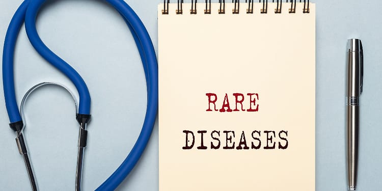 A notebook with the phrase "RARE DISEASES" written in large letters, accompanied by a blue stethoscope and a silver pen on a light background.