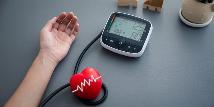 A human arm with hand outstretched next to a digital blood pressure monitor, showing readings of 125 mmHg and 80 mmHg, plus a red heart-shaped device with a pulse line, decorated with small wooden houses and a flower pot in the background.