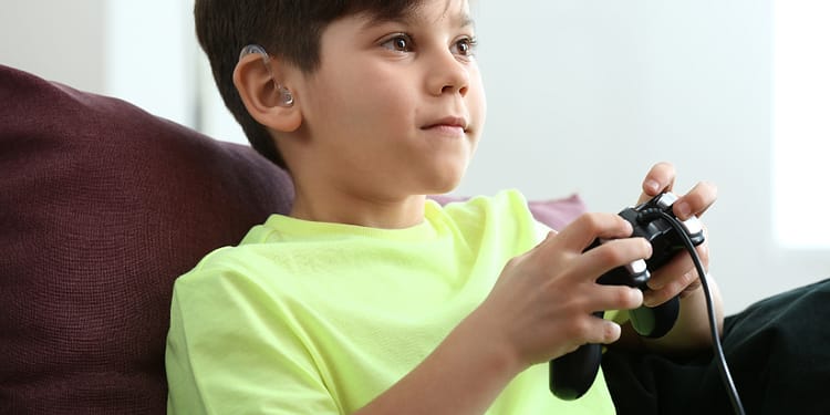 Un niño sentado en un sofá, usando un controlador de videojuegos y vistiendo una camiseta amarilla brillante, con un audífono visible en su oído izquierdo, mostrando una expresión concentrada mientras juega.