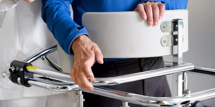 A caregiver holds a person wearing a blue shirt while the person holds onto a harness on a mobility assist device.