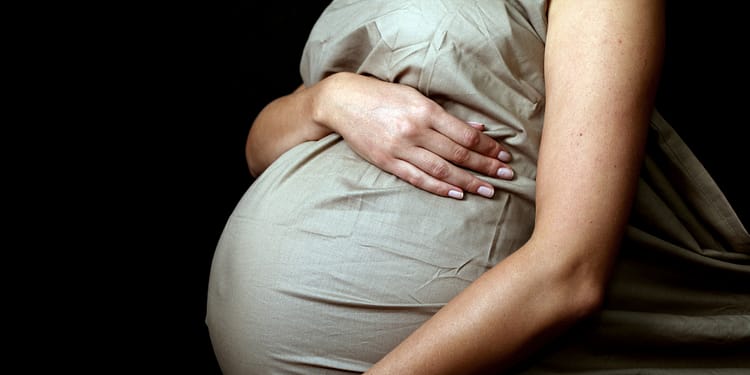 Belly of a pregnant woman covered with a light fabric, with hands resting on her belly, on a dark background.