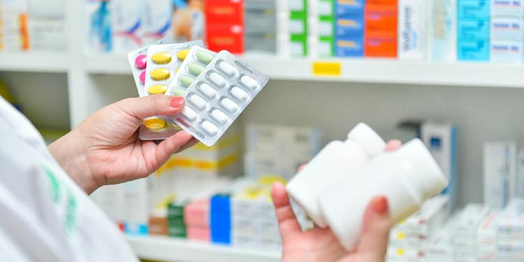A person holds three blister packs of colored pills and two white bottles of medication, in a pharmacy setting with shelves full of boxes of different colored medications in the background.