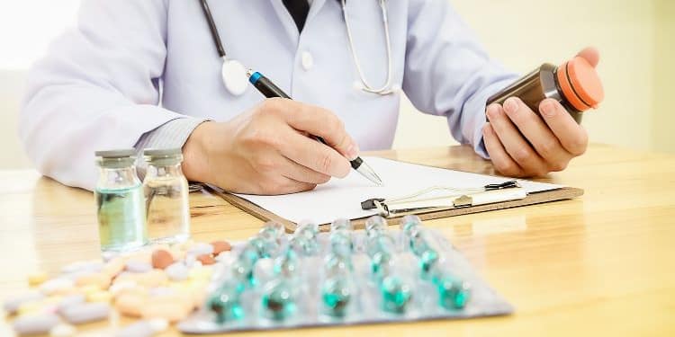 A doctor with a stethoscope dressed in a white coat holds a bottle of medication in one hand and writes on a form with the other, while vials and a variety of pills lie on the table.