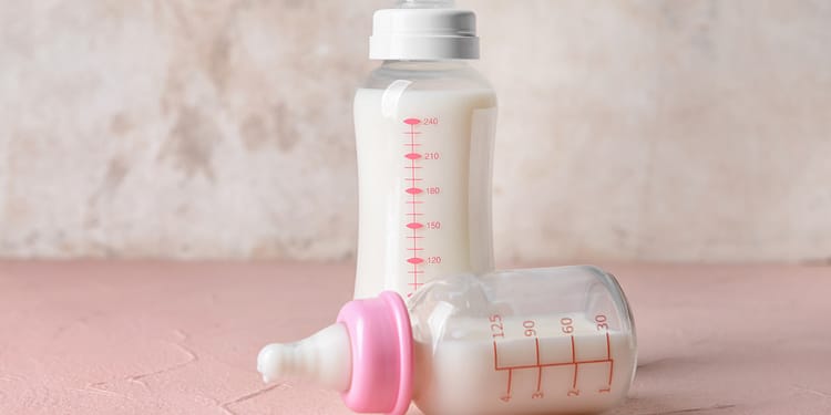 Plastic milk bottles, one white with a white cap and red scale, and one transparent with a pink cap and red scale, on a pink surface.
