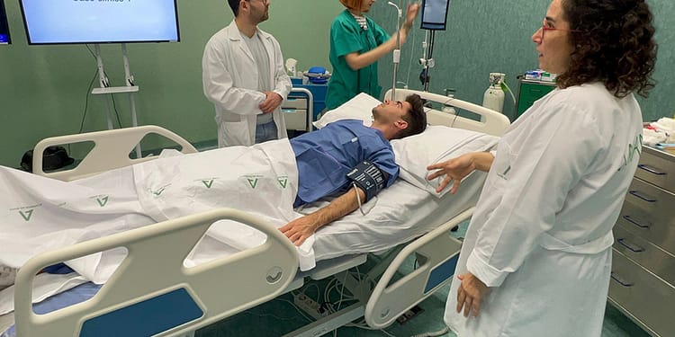 Two doctors and a nurse interact with a patient in a hospital bed. A monitor displays "Clinical Case 1." The patient is semi-reclined with a blood pressure cuff.