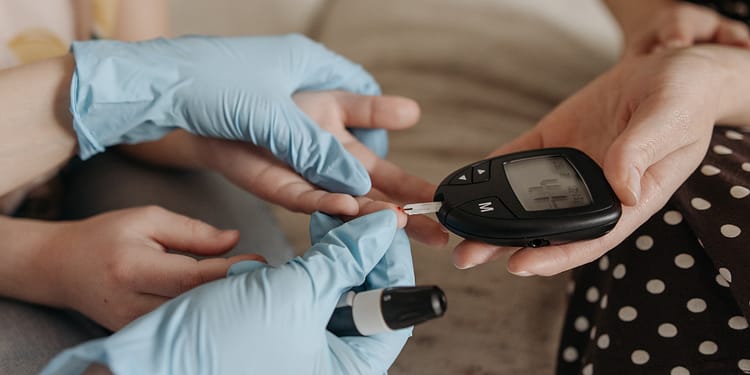 A person wearing latex gloves is using a blood glucose monitoring device on another hand, while preparing to apply a blood sample.