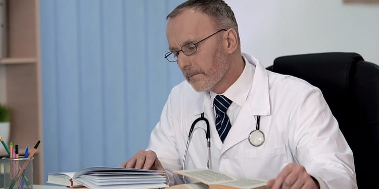 A doctor in a white coat and glasses, with a stethoscope around his neck, studying several open books on a desk, showing a concentrated expression, against a bookshelf and a light wall.