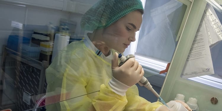 Mujer en un laboratorio, vestida con una bata amarilla y cofia, utilizando un micropipeta para transferir líquido en un frasco.