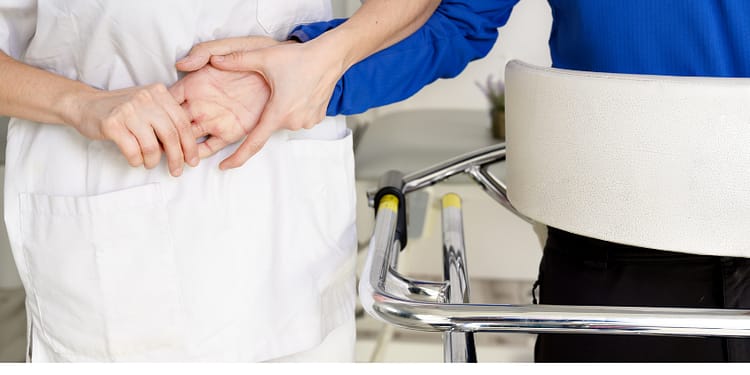 A person in a white uniform holds the hand of another person in a blue T-shirt, in an environment that appears to be a clinic or healthcare facility. A device for assistance can be seen in the background.