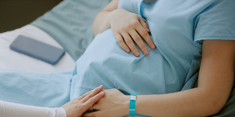 A pregnant person on a bed, with one hand on her abdomen and another hand touching the pregnant person's hand. Both are wearing comfortable clothes and there is a mobile phone on the bed.