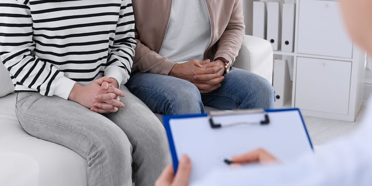 A person in a striped sweater and another in a jacket sit on a sofa, while someone else, out of focus, is taking notes in a folder.