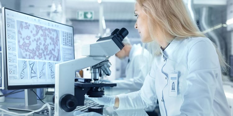 Mujer de laboratorio mirando a través de un microscopio, con una pantalla de computadora que muestra gráficos y datos científicos.