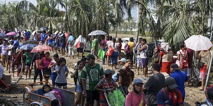Group of people in line, many with umbrellas, in a tropical setting with palm trees, possibly waiting for help or resources.
