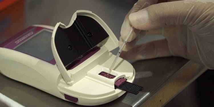 A medical monitoring device with an open lid, in which a gloved hand inserts a pipette into the front of the device.
