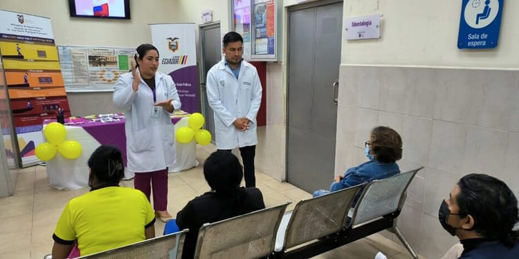 Two health professionals, dressed in white coats, are speaking in front of a group of people sitting in a waiting room. There are yellow balloons decorating the table in the background and informative posters on the walls.