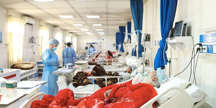 Hospital room with several patients in beds, medical staff in blue suits and masks, and medical equipment visible in a well-lit environment.